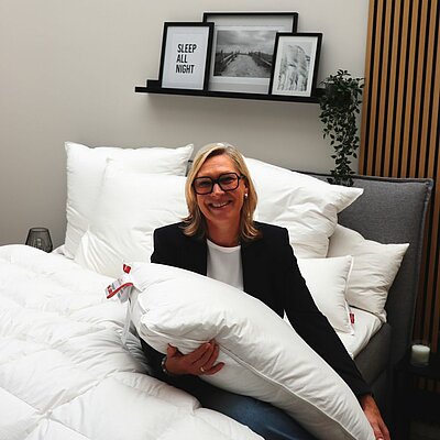 Die neue Geschäftsführerin sitzt entspannt auf einem Bett, hält ein Kissen und lächelt freundlich in die Kamera.
