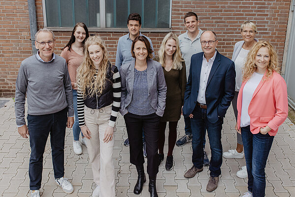 Gruppenfoto von zehn Mitarbeiter*innen, die im Freien vor einer Backsteinwand stehen.