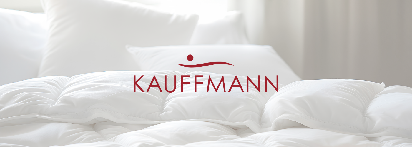 Logo "Kauffmann". Im Hintergrund eine weiße Bettdecke und ein Kopfkissen.