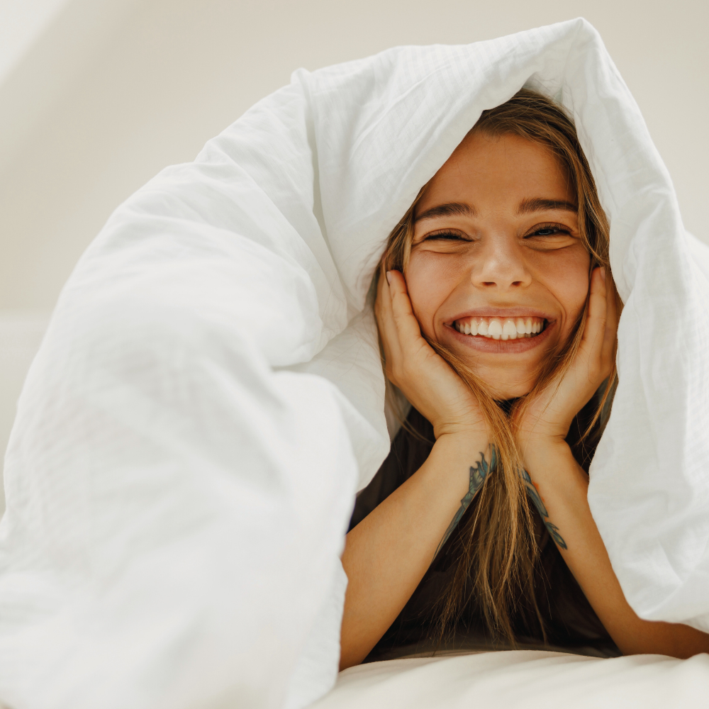 Eine Frau liegt unter einer weißen Bettdecke und lächelt fröhlich in die Kamera.